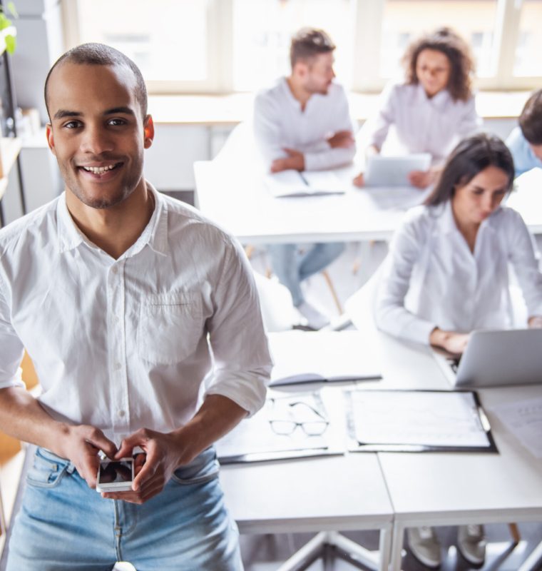 Successful young business people are working in office. Guy in the foreground is using a smart phone,  looking at camera and smiling